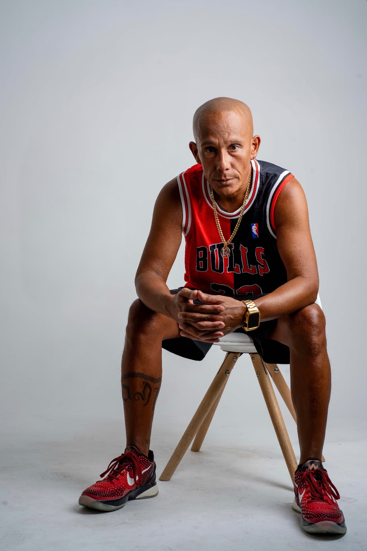 MC Rony mit Glatze sitzt auf einem Hocker vor hellem Hintergrund. Er trägt ein rot-schwarzes Chicago Bulls Trikot, eine Goldkette und rote Turnschuhe.