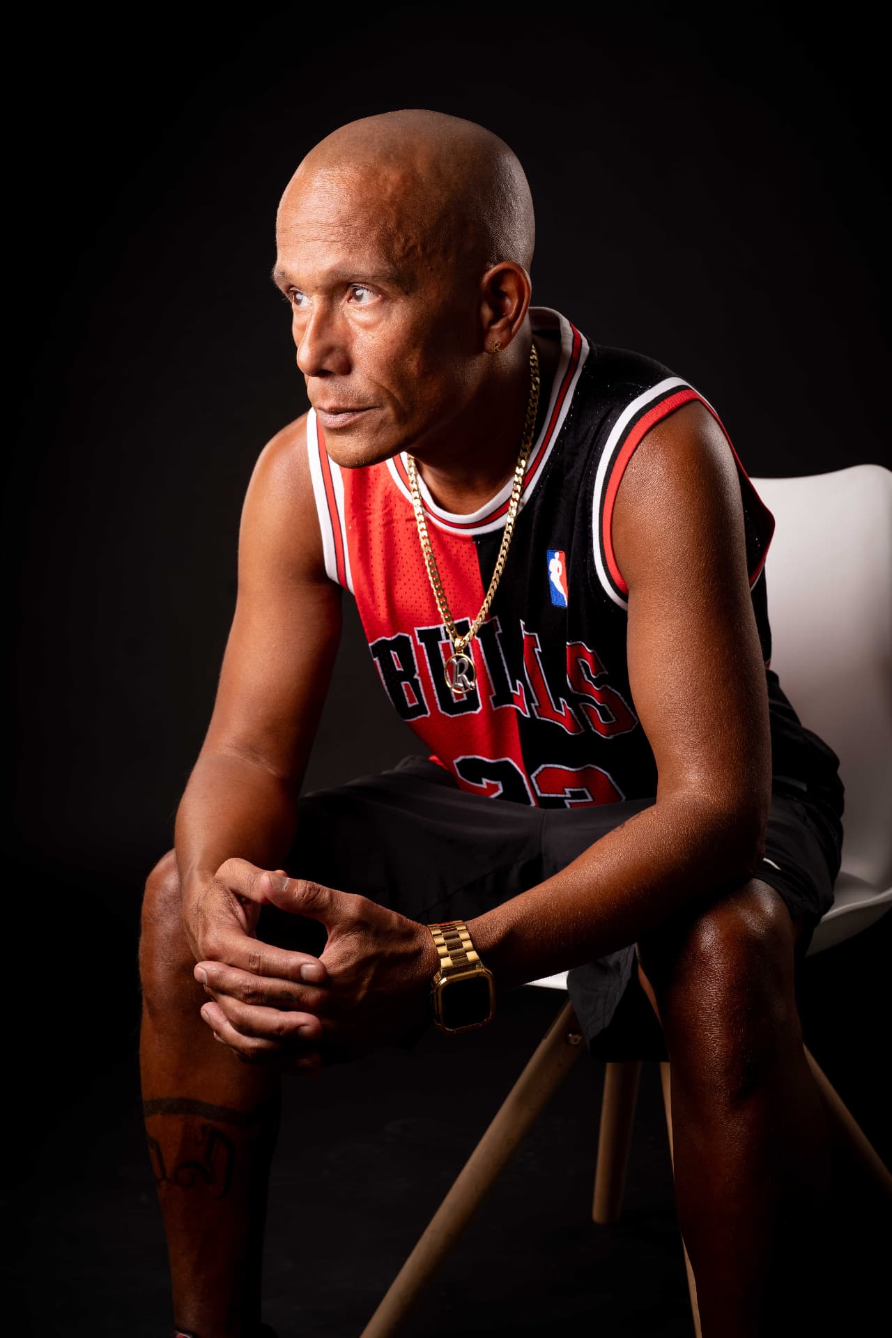 Porträt von MC Rony, sitzend auf einem weissen Stuhl, trägt ein rot-schwarzes Bulls-Jersey, Goldkette und Uhr vor schwarzem Hintergrund, nachdenklicher Blick.