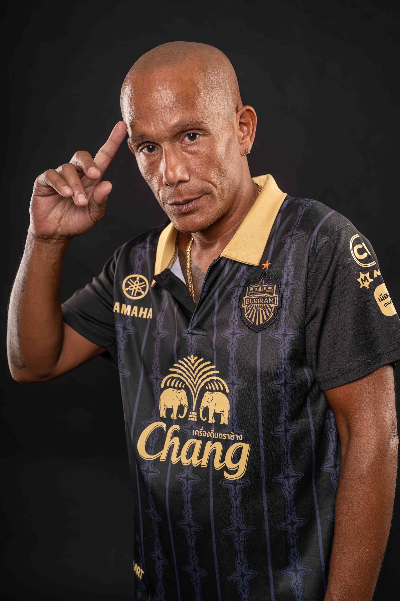 Porträt von MC Rony mit Glatze und Sporttrikot vor dunklem Hintergrund, ein Finger an der Schläfe, ernster Blick.