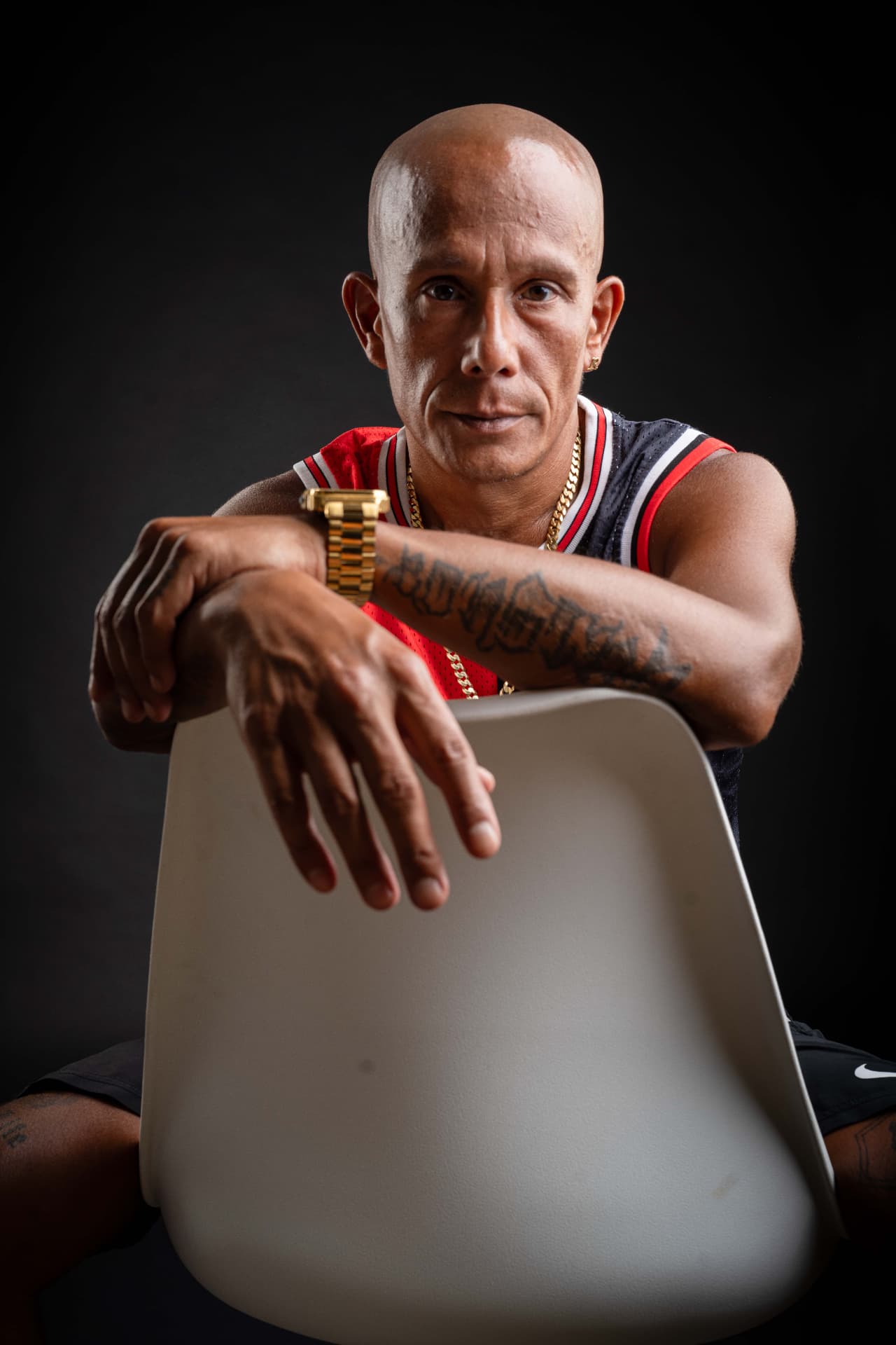 MC Rony, ein Mann mit Glatze und Tätowierung, sitzt verkehrt herum auf einem weissen Stuhl. Er trägt ein Basketball-Trikot, eine Goldkette und eine Golduhr.