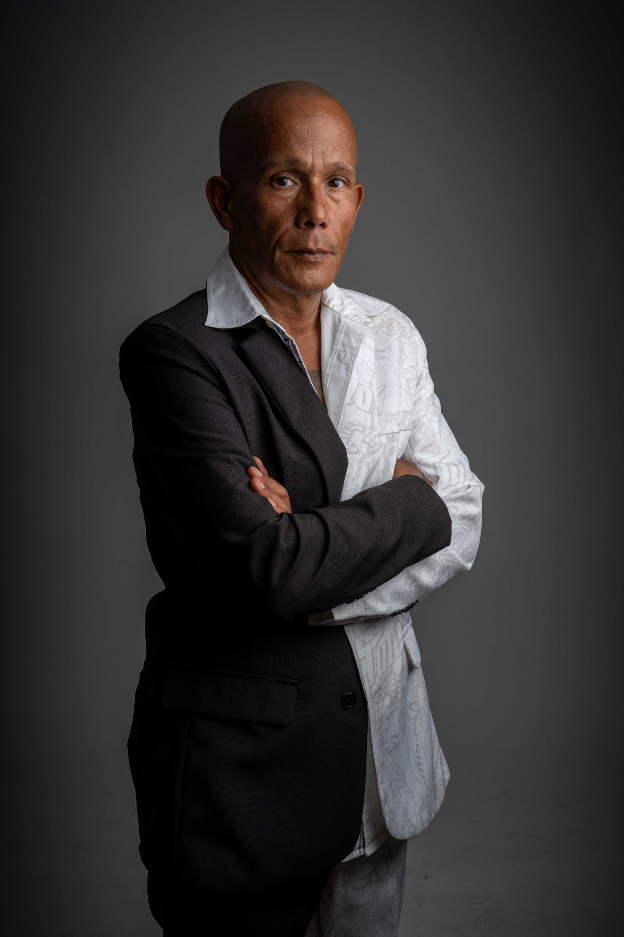Portraet von MC Rony mit Glatze, verschraenkten Armen und einem zweifarbigen Sakko (schwarz/weiss gemustert) vor grauem Hintergrund.